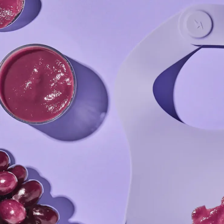 A modern, minimalist silicone bib with a spill pocket, placed on a pastel purple background next to a smoothie and fresh grapes.
