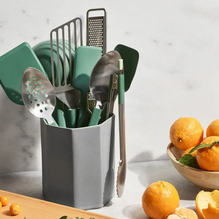 A stylish kitchen utensil set with green and stainless steel tools stored in a sleek gray holder, surrounded by fresh oranges and a marble countertop.