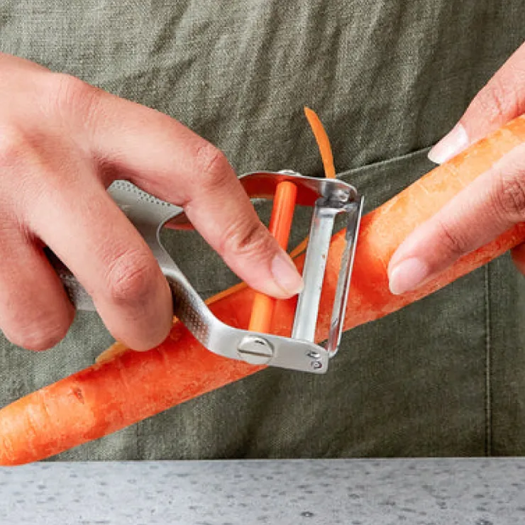 Close-up of hands peeling a fresh carrot with a stainless steel peeler against a neutral fabric backdrop and a marble countertop.